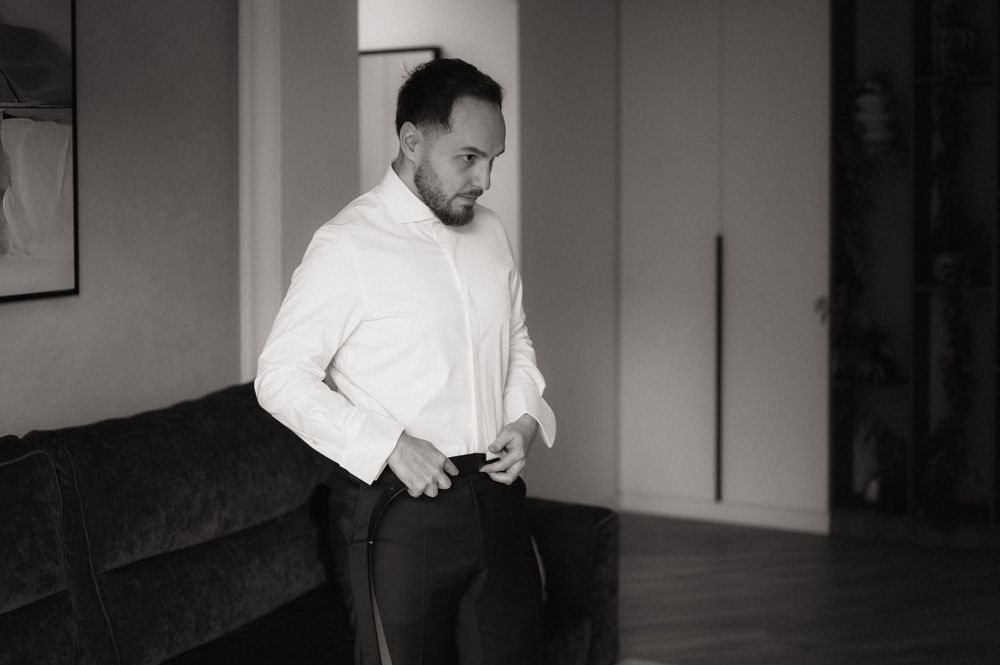 Black and white photo of a man in a white dress shirt buttoning up while getting ready indoors.