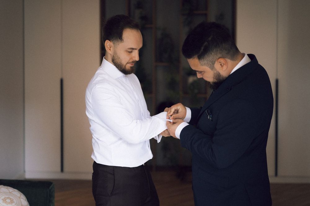 Two men in formal attire assist each other with cufflinks while getting ready for a wedding ceremony.
