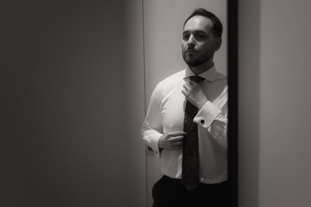 Black and white photo of a man in a white dress shirt adjusting his tie while looking in a mirror.