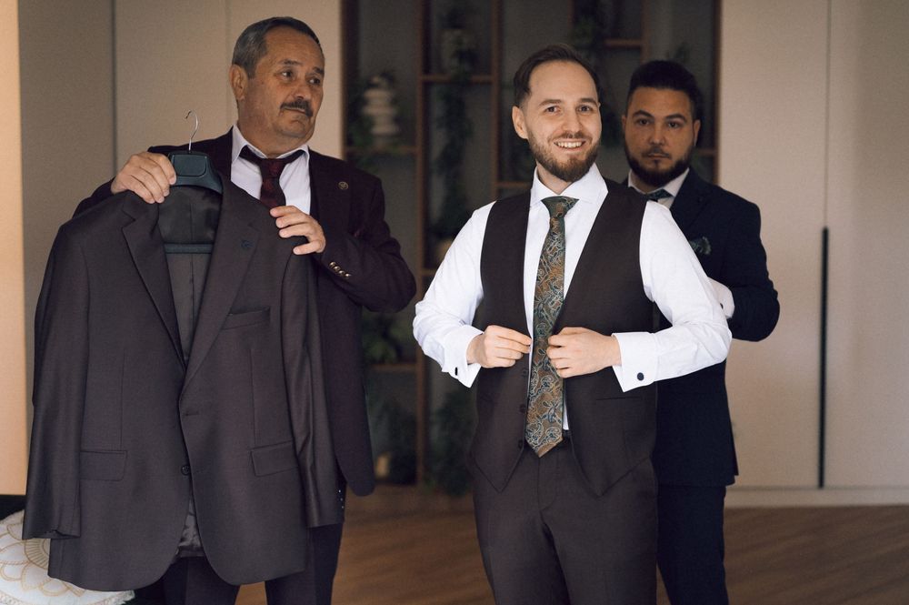 Three men in formal suits preparing for a wedding, with one holding a jacket on a hanger indoors.