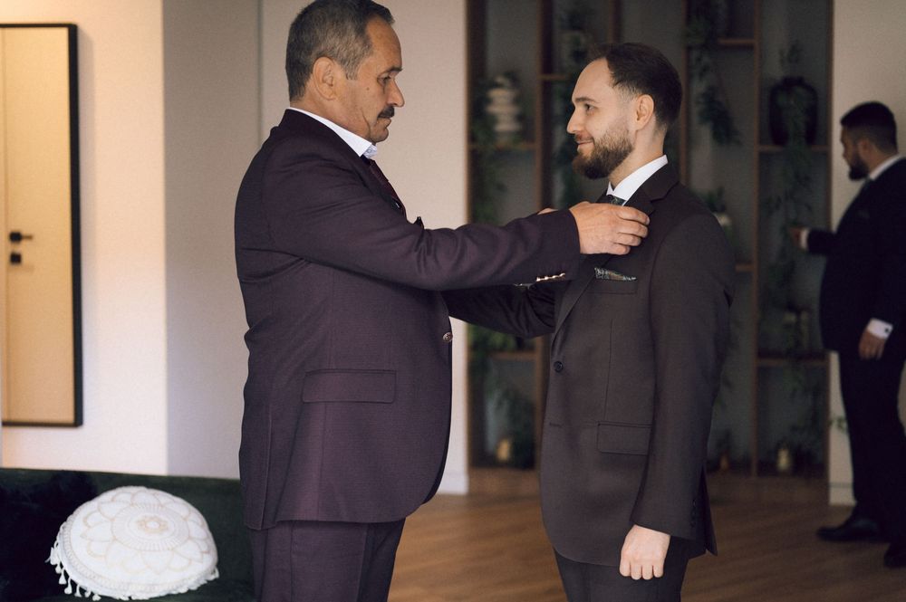Two men in dark suits, one adjusting the other's collar in an elegant room during a formal occasion.