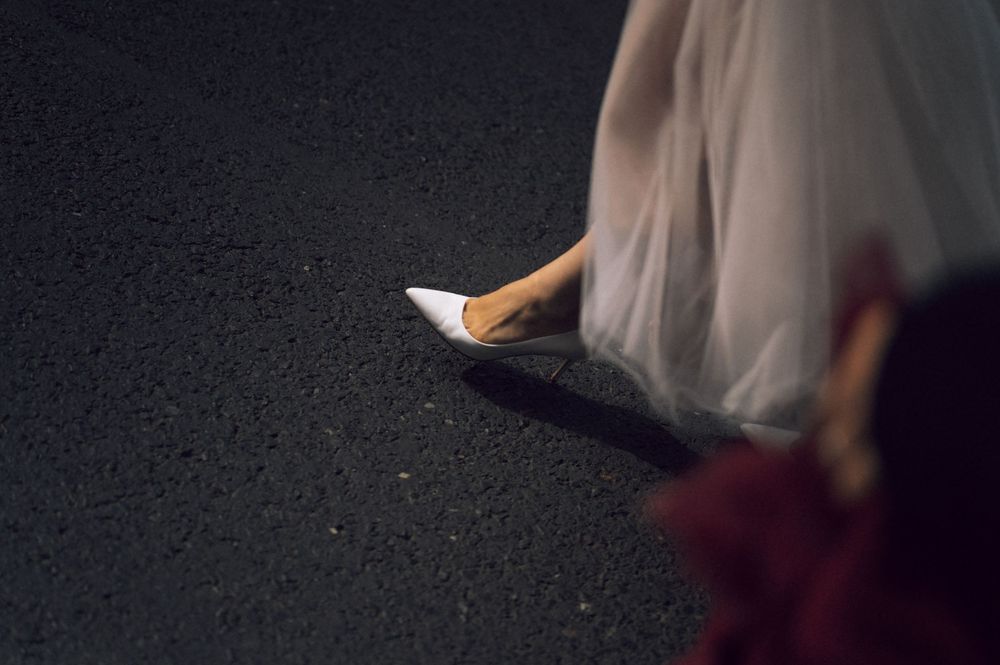 Close-up of a bride's white pointed-toe heels and flowing tulle wedding dress hem on dark pavement.