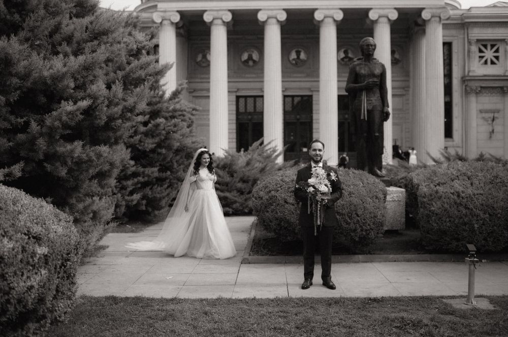 Black and white wedding photo of bride in white gown and groom in suit posing before a columned building with statue.