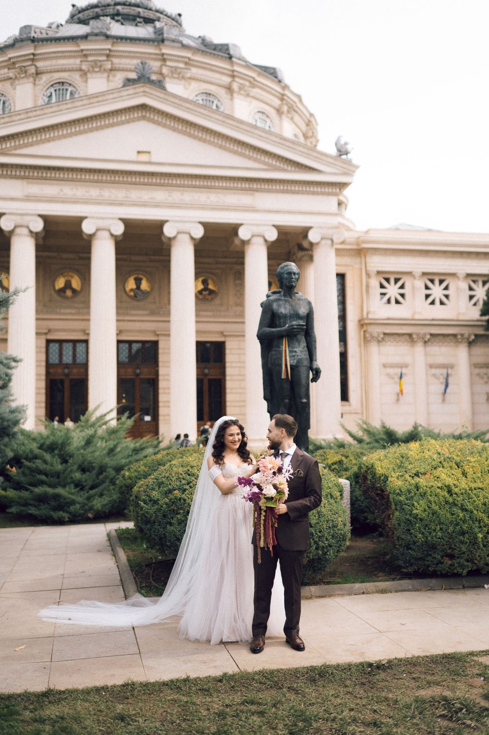 Bride and groom pose in front of the Romanian Athenaeum in Bucharest, bride holding a colorful bouquet.