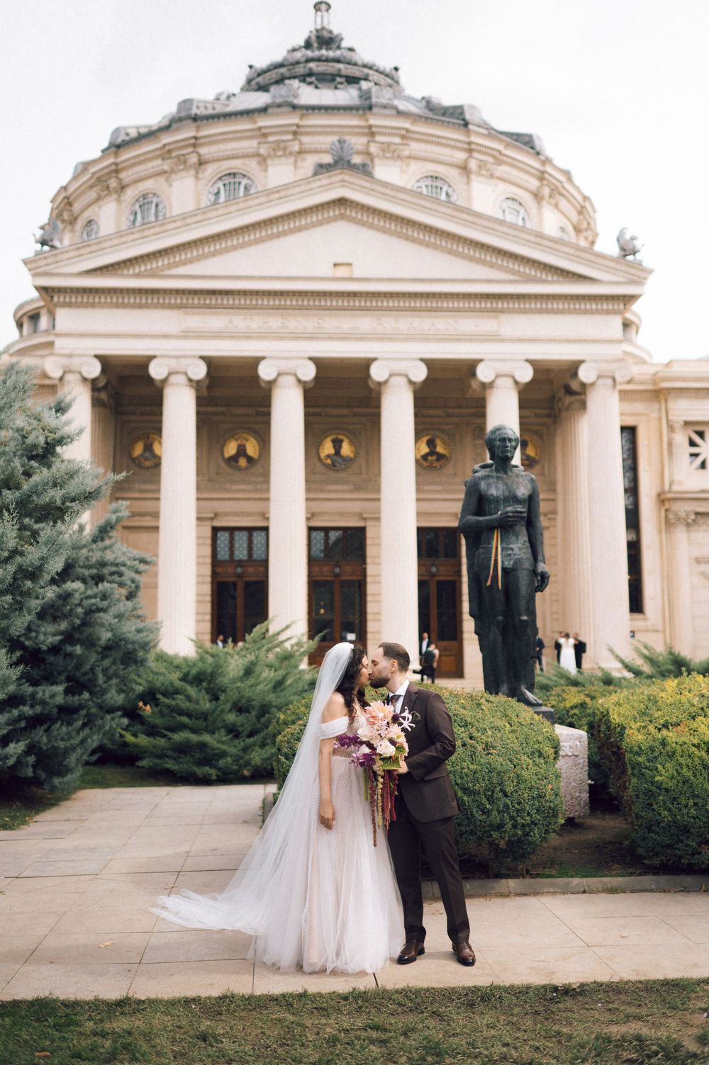 Bride and groom kissing in front of the Romanian Athenaeum neoclassical building with columns and statue.