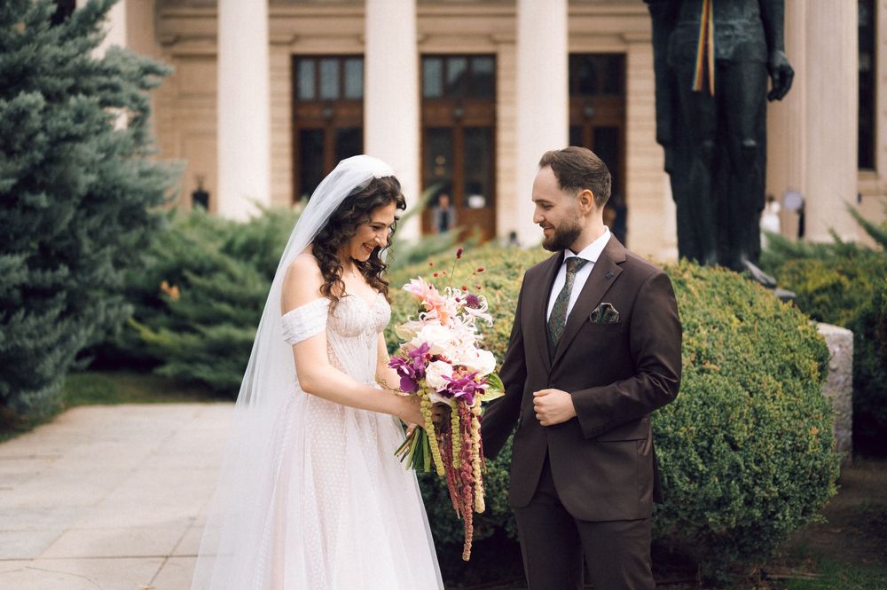 Bride in white gown holding bouquet stands with groom in brown suit outside elegant building.