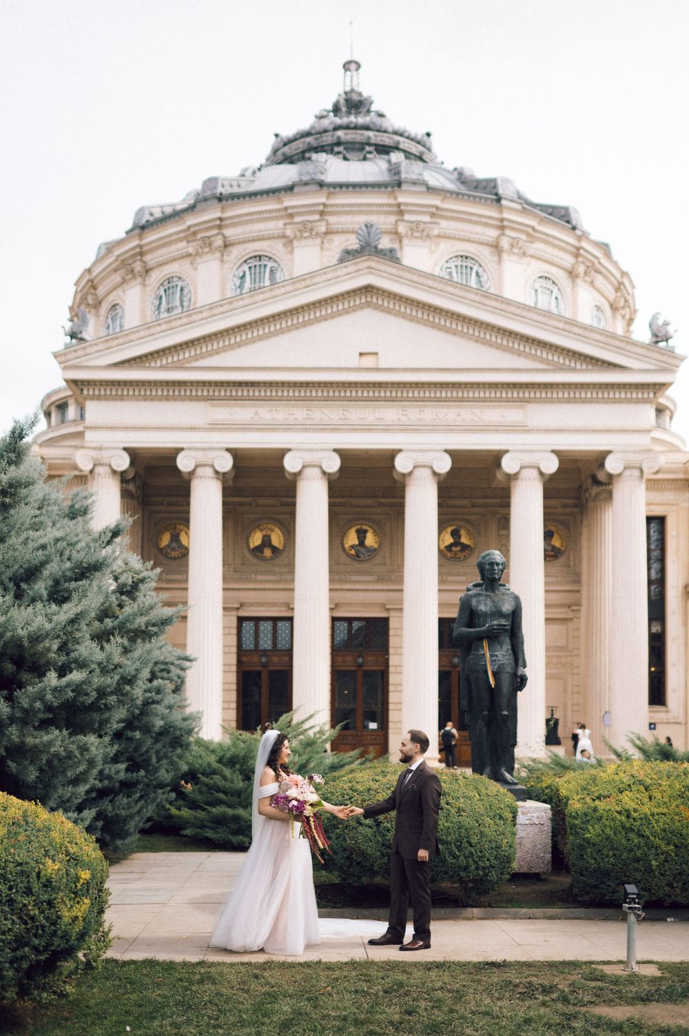Bride and groom hold hands before the Romanian Athenaeum neoclassical building with columns and statue.