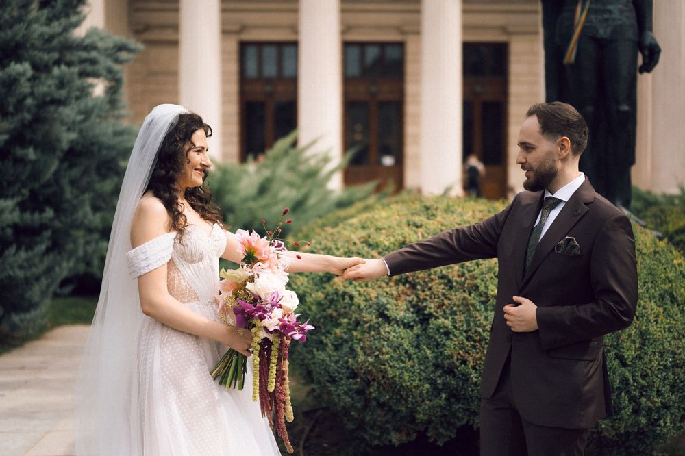 Bride and groom holding hands outdoors, bride in white gown with bouquet, groom in dark suit, elegant building backdrop.