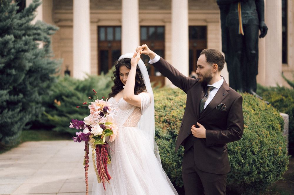 Groom twirling bride in white gown and veil, holding colorful bouquet, posing outdoors near classical building.