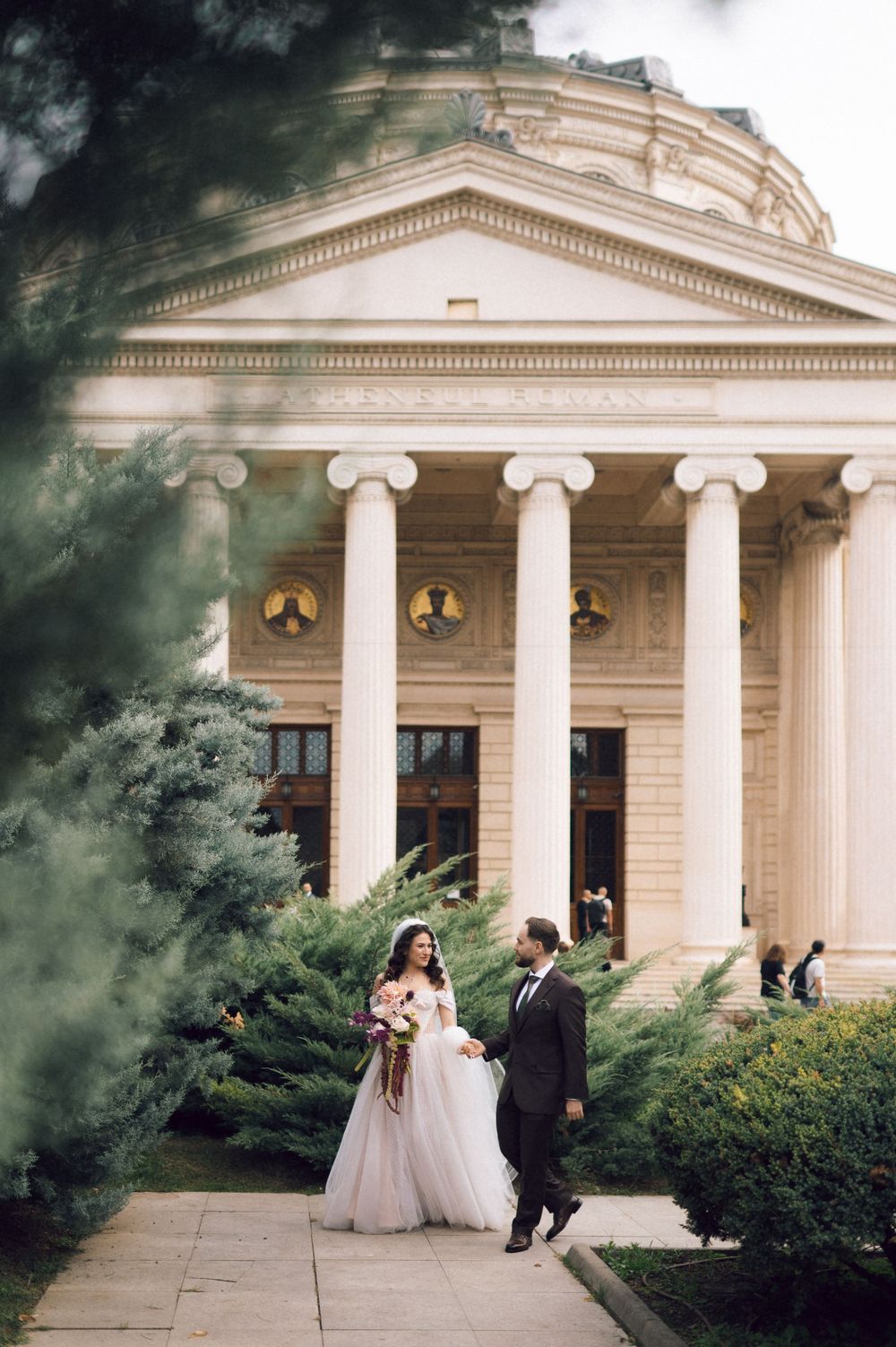 Bride and groom walking hand in hand in front of a grand neoclassical building with tall columns.