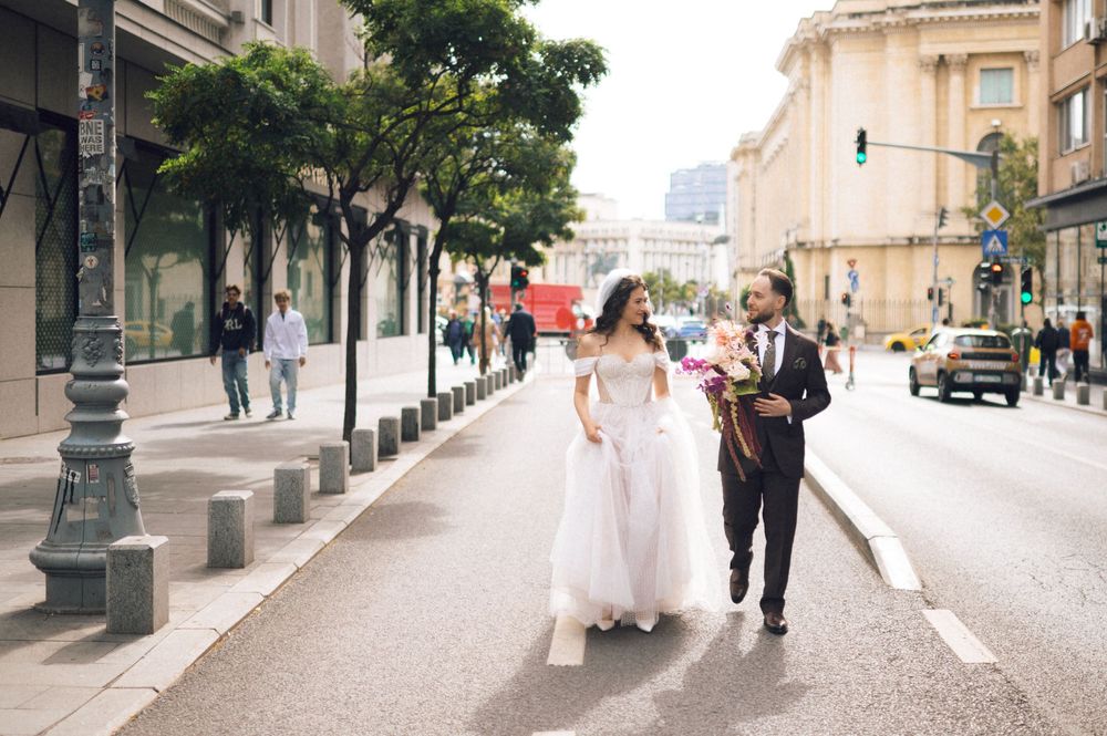 Bride and groom walking hand in hand across a city street, bride in white gown, groom in dark suit with bouquet.