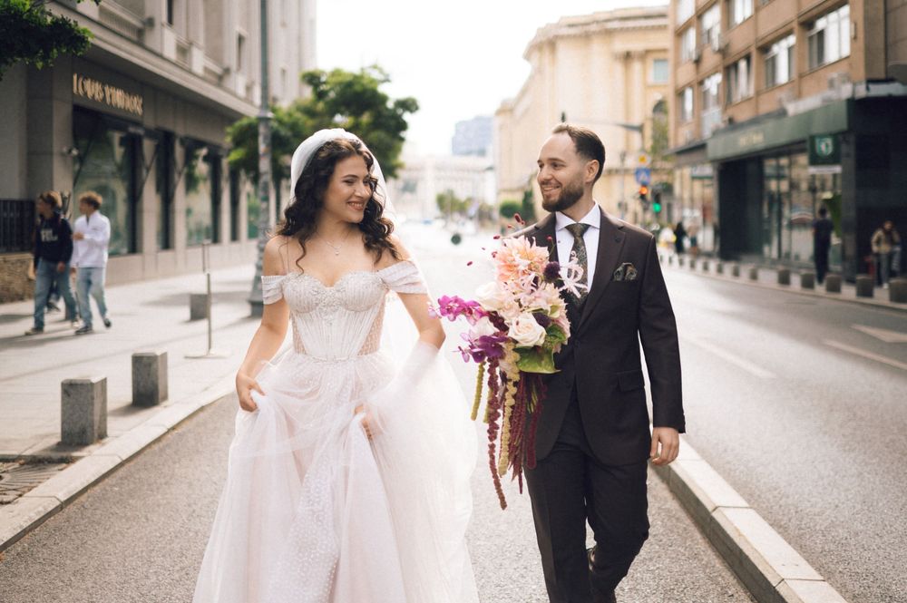 Bride and groom walking on a city street, bride in white gown holding colorful bouquet, groom in dark suit.