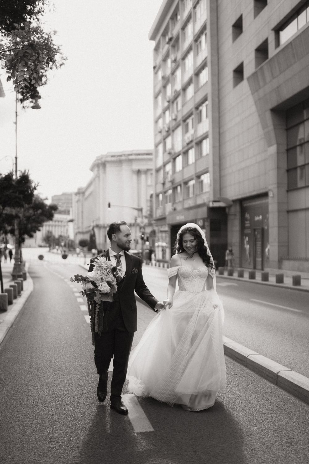 Black and white photo of a bride and groom holding hands walking down a city street, bride in a flowing gown.