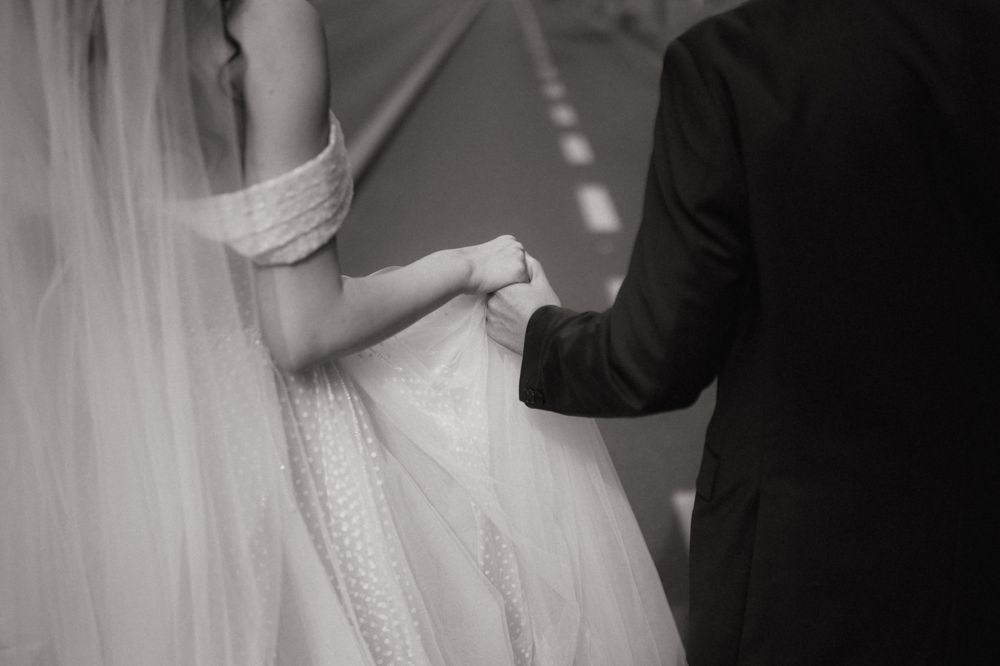Black and white photo of bride and groom holding hands, showcasing wedding dress and veil.