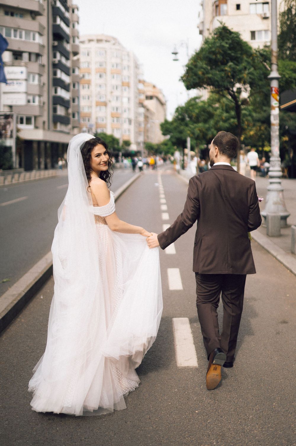 Bride in white gown and veil holds grooms hand walking across city street, smiling back at camera.