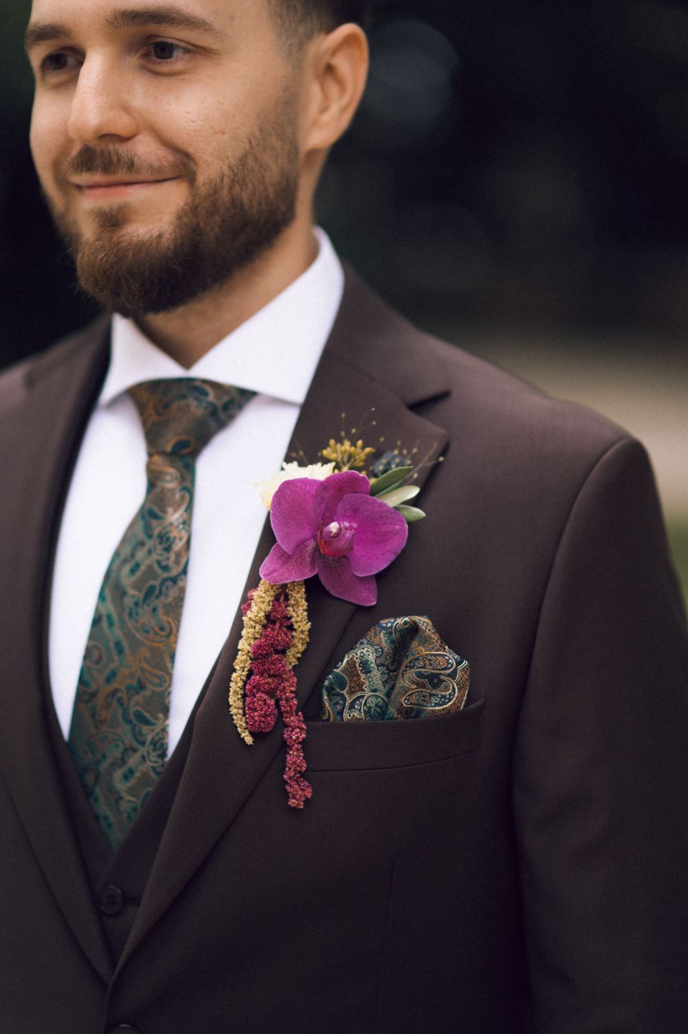 Groom in dark brown suit with floral tie, pocket square, and vibrant pink orchid boutonniere at a wedding.