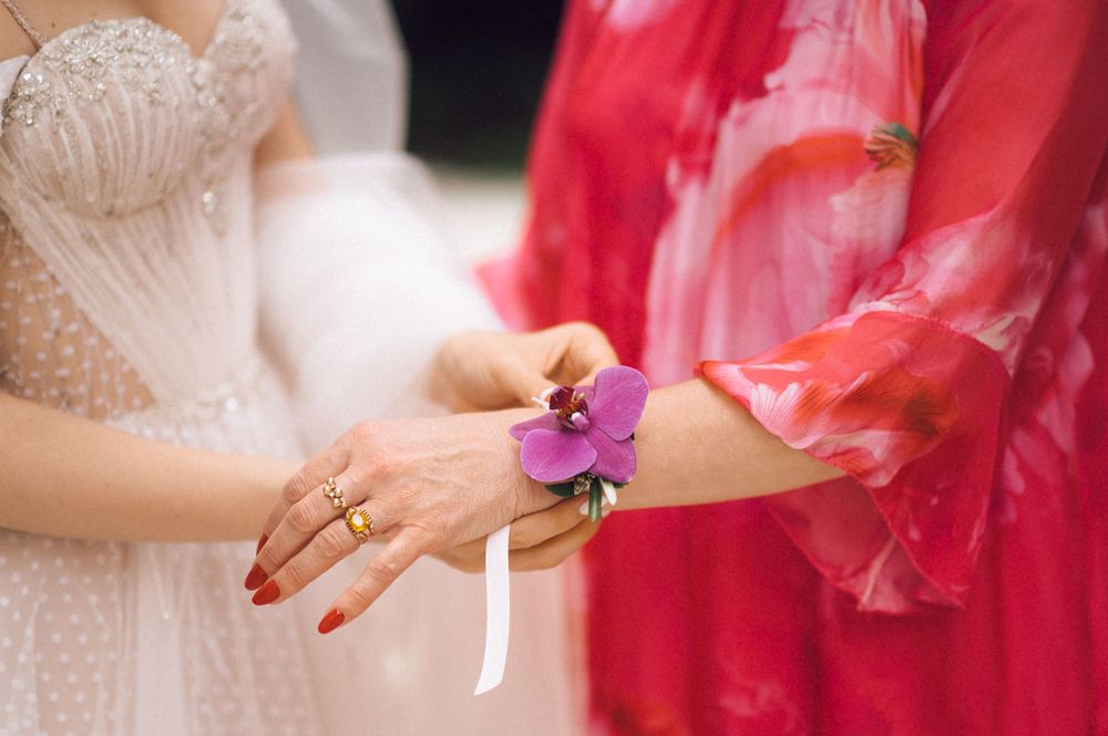 Bride in white polka dot dress attaching purple orchid wrist corsage on woman in red floral dress.