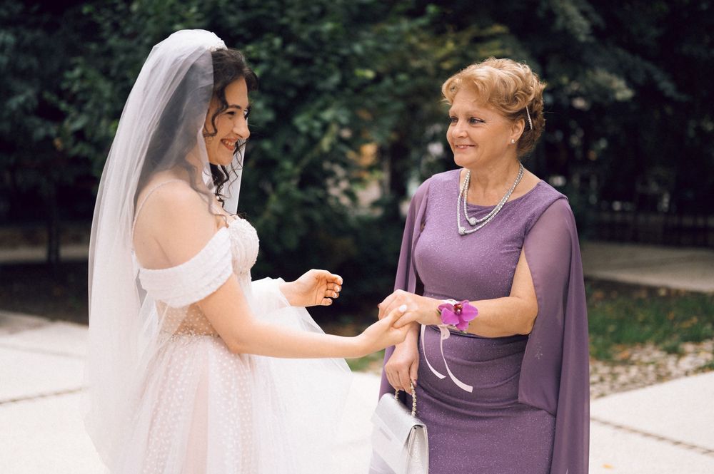 Bride in white gown and veil shares a joyful moment with older woman in purple dress outdoors.