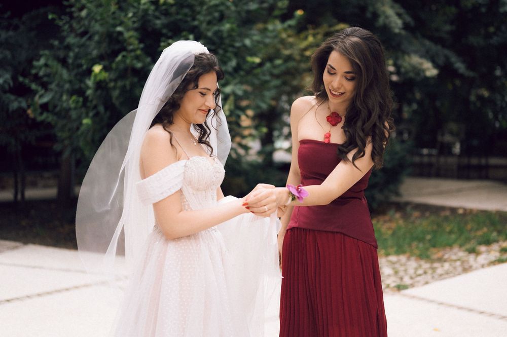 Bride in white gown and veil with bridesmaid in burgundy dress examining wedding details outdoors.