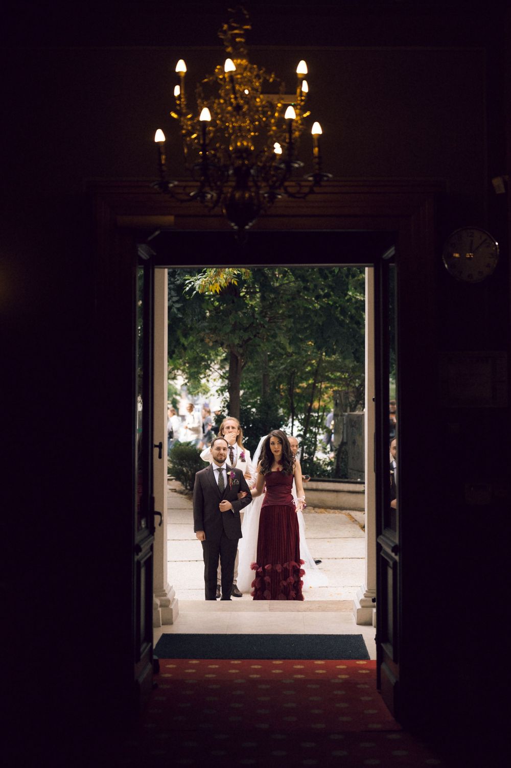 Couple in formal attire standing in elegant doorway with chandelier overhead and lush greenery outside.