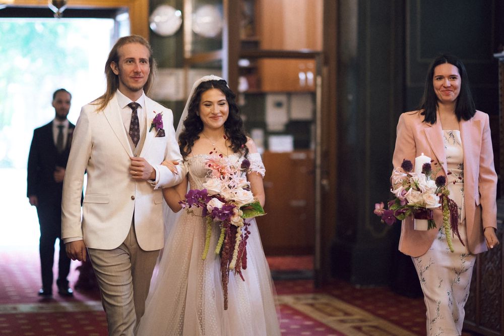 Wedding party walks down aisle, bride in white gown holding purple bouquet, groom in beige suit, bridesmaid in pink.