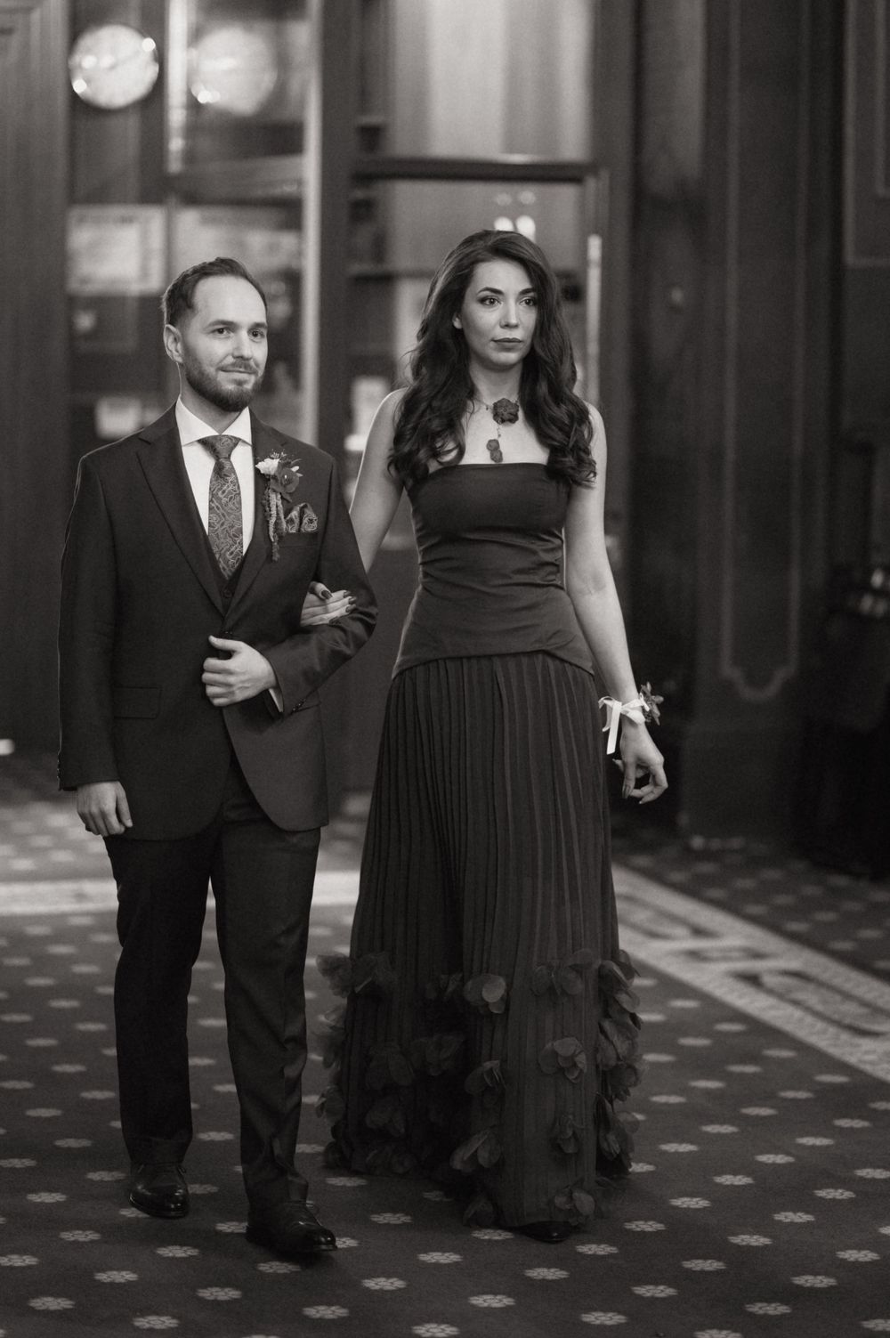 Elegant couple in formal attire walking together, man in suit with boutonniere, woman in long black dress, black and white photo.