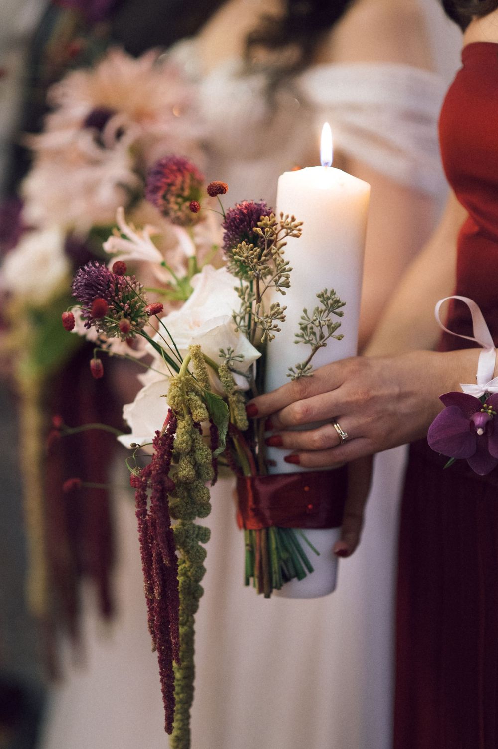 Bridesmaids holding bouquets of purple and white flowers with a lit white pillar candle at a wedding ceremony.