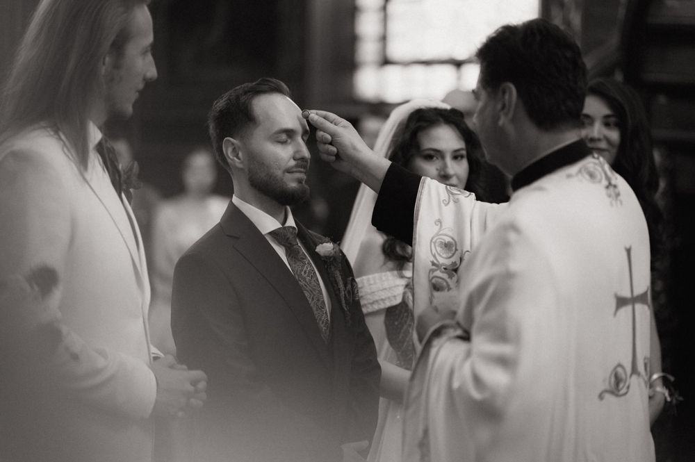 Black and white photo of a groom receiving a blessing from a priest during a religious wedding ceremony.
