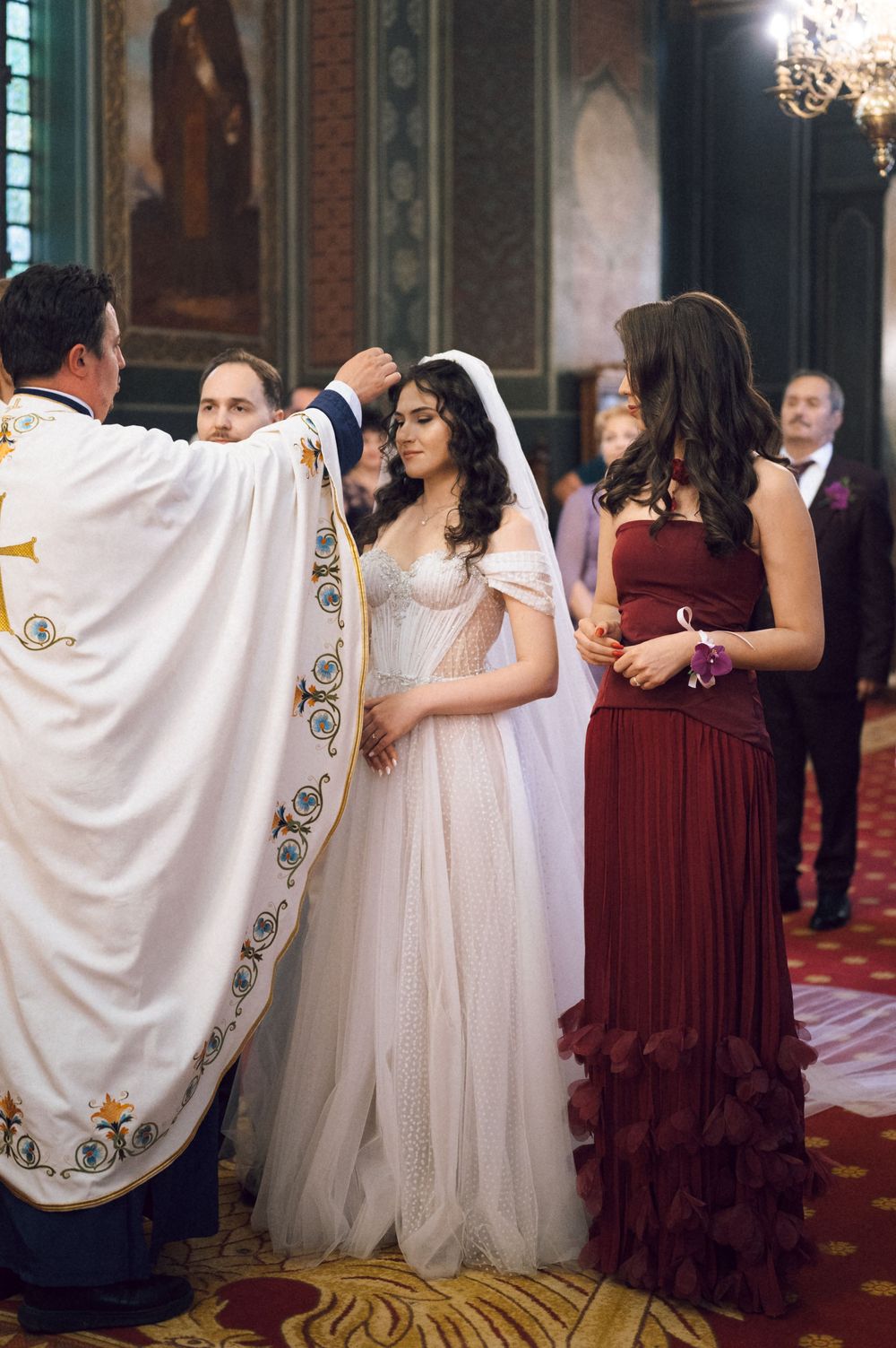Priest blessing bride in white wedding gown during church ceremony with bridesmaid in burgundy dress.