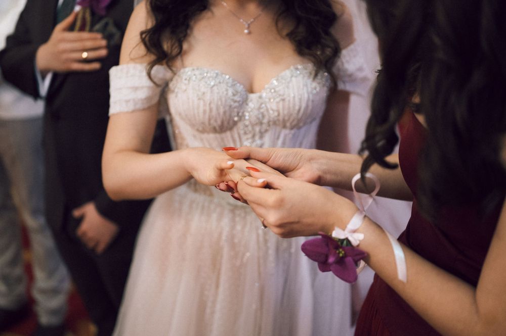 Bride in white lace wedding gown receives ring from bridesmaid in burgundy dress during wedding ceremony.
