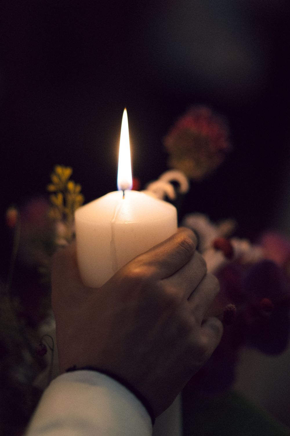 Hands holding a glowing white pillar candle with flowers in the background during a wedding ceremony.