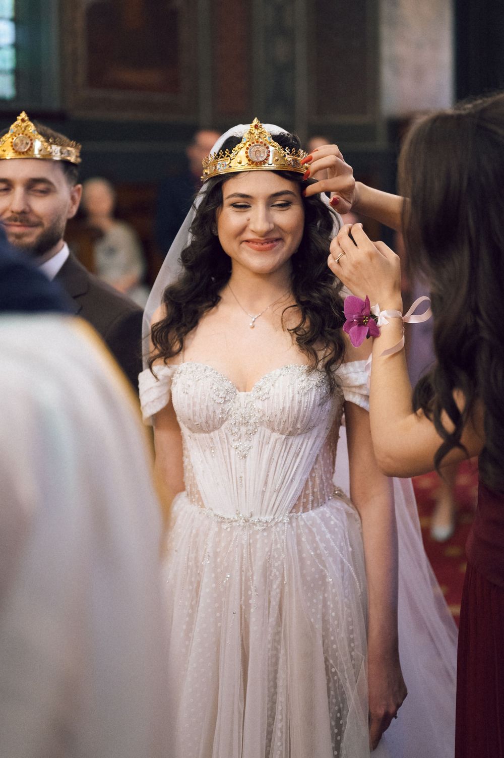Smiling bride in a white gown receiving a gold crown during an Orthodox wedding ceremony.
