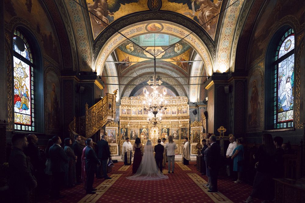 Elegant Orthodox church wedding ceremony with ornate golden iconostasis, stained glass windows, and guests lining the red carpet aisle.