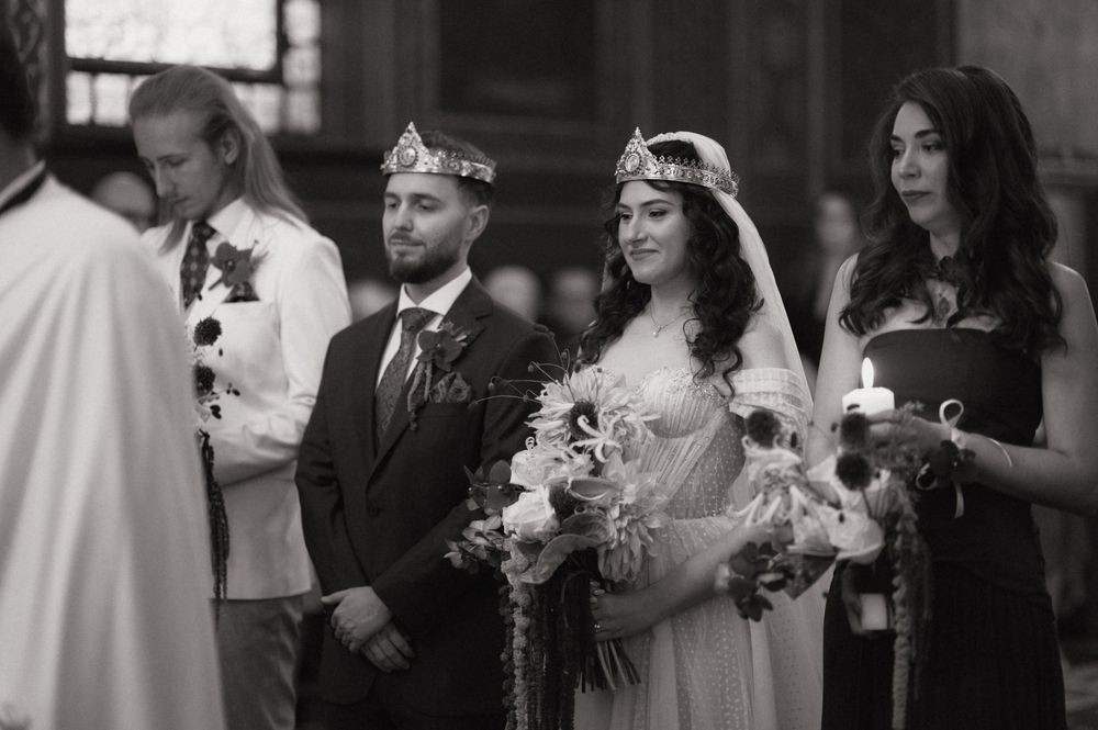 Black and white wedding photo of bride and groom wearing crowns at church ceremony with attendants holding candles.