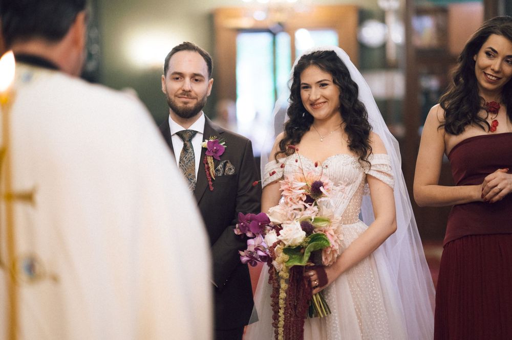 Bride and groom smile during wedding ceremony, bride holding purple floral bouquet in elegant white gown with veil.