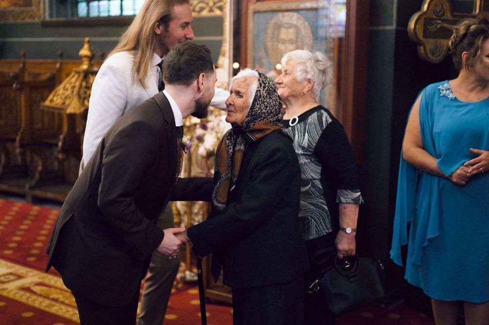 Elderly woman greeted by young man in suit inside ornate Orthodox church with icons and gold decor.