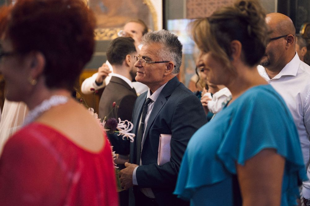 Elegantly dressed guests mingle at an indoor social event, with a man in a suit holding documents in the foreground.