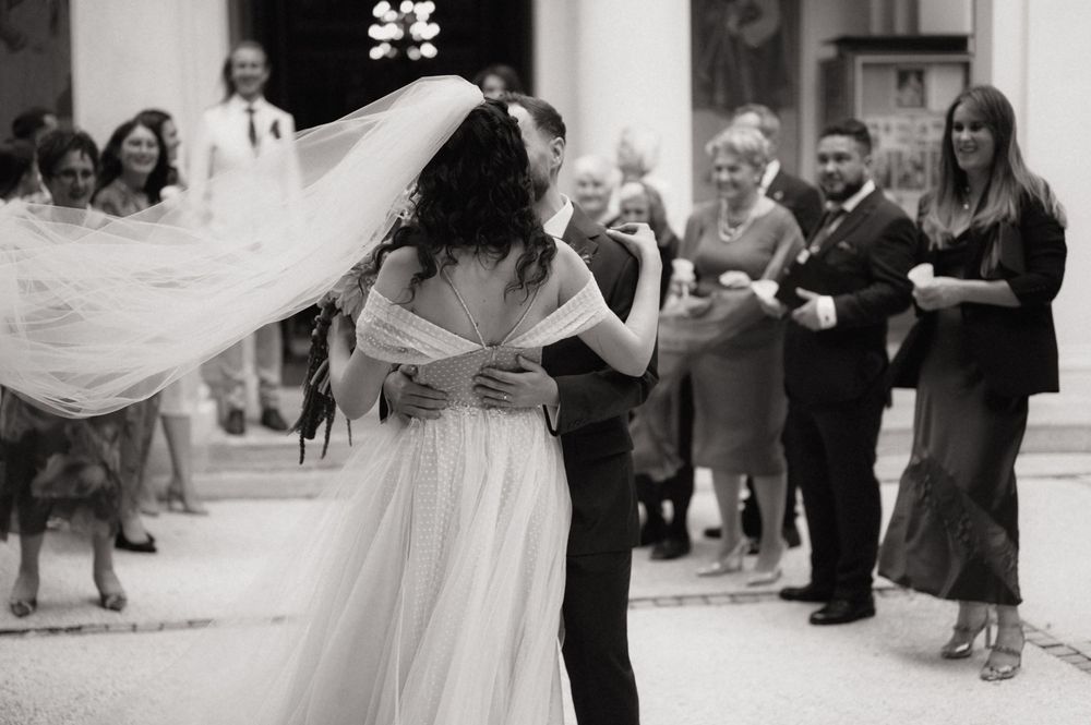 Black and white photo of bride and groom sharing first dance at wedding ceremony outdoors with guests watching.