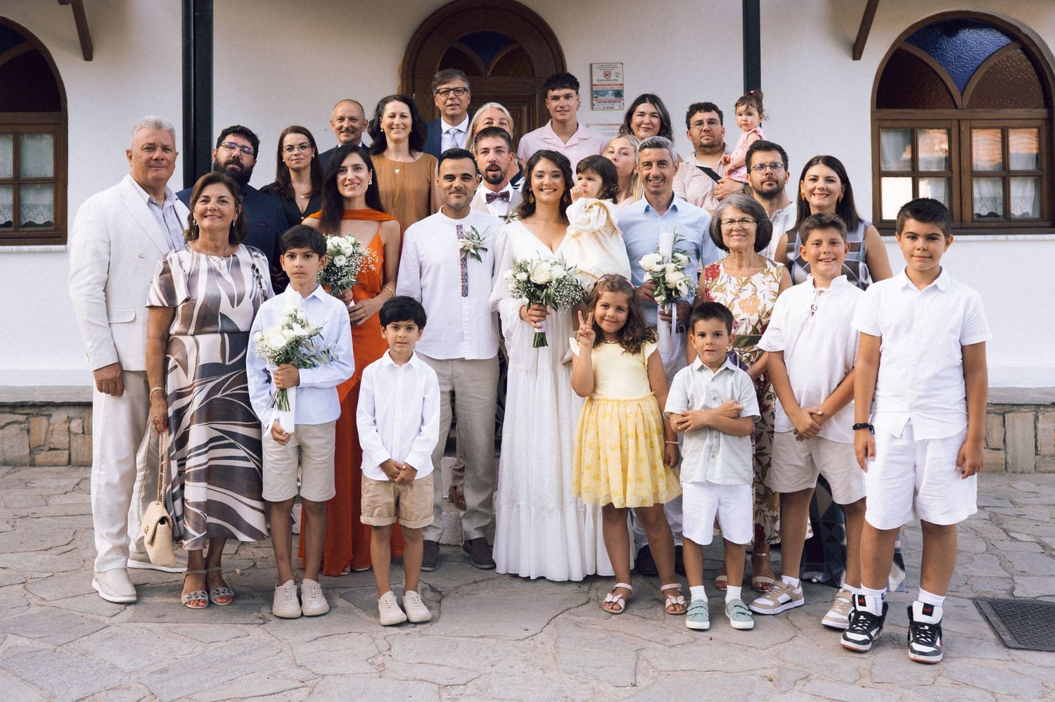 Large family group photo at a wedding ceremony outdoors, with bride and groom in white surrounded by guests.