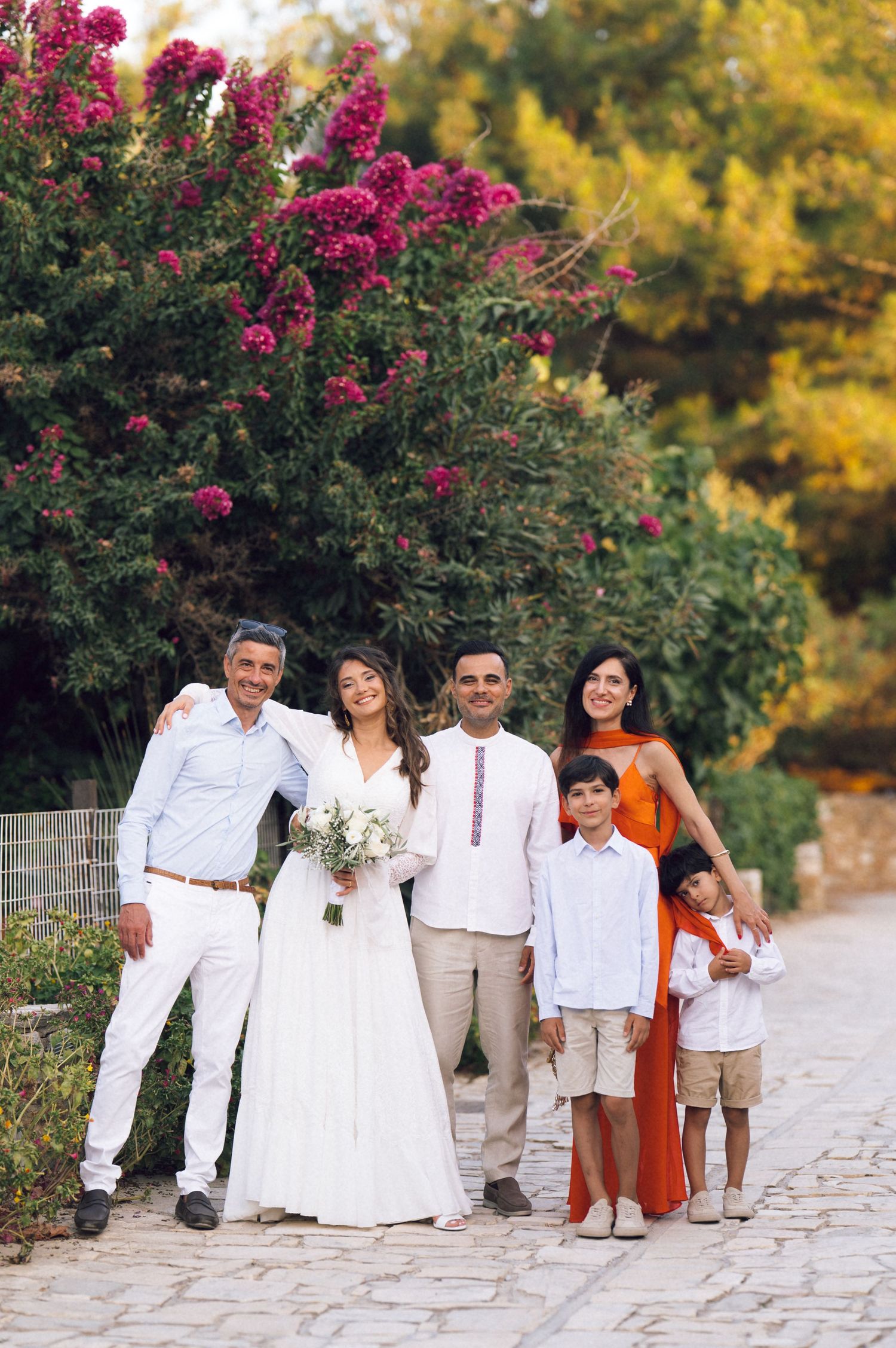 Family group photo at a wedding outdoors, bride in white gown holding bouquet, surrounded by guests near blooming trees.