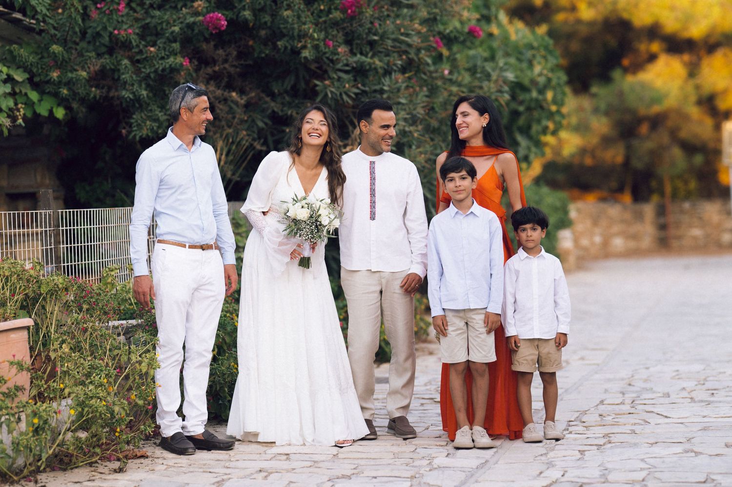 Wedding party of six poses on a stone path, bride in white gown holding bouquet, surrounded by family in elegant attire.