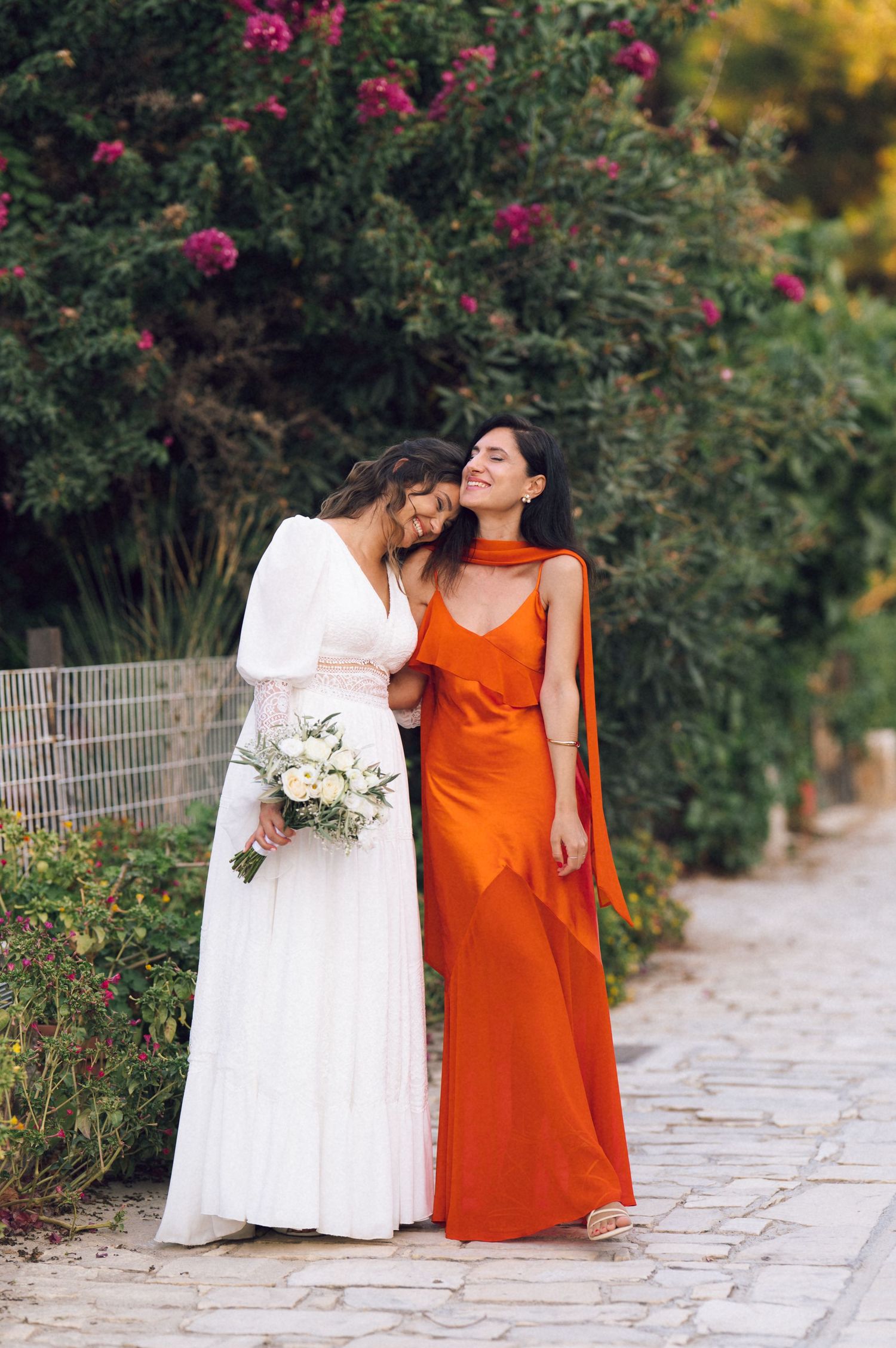 Bride in white gown with bouquet and bridesmaid in orange satin dress laughing together on garden path.