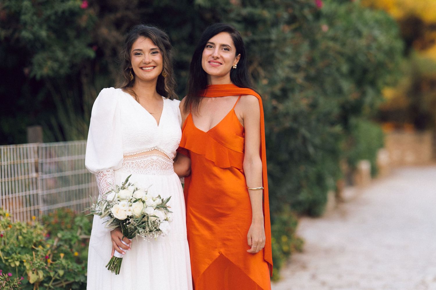 Bride in white gown holding bouquet poses with bridesmaid in orange dress outdoors.