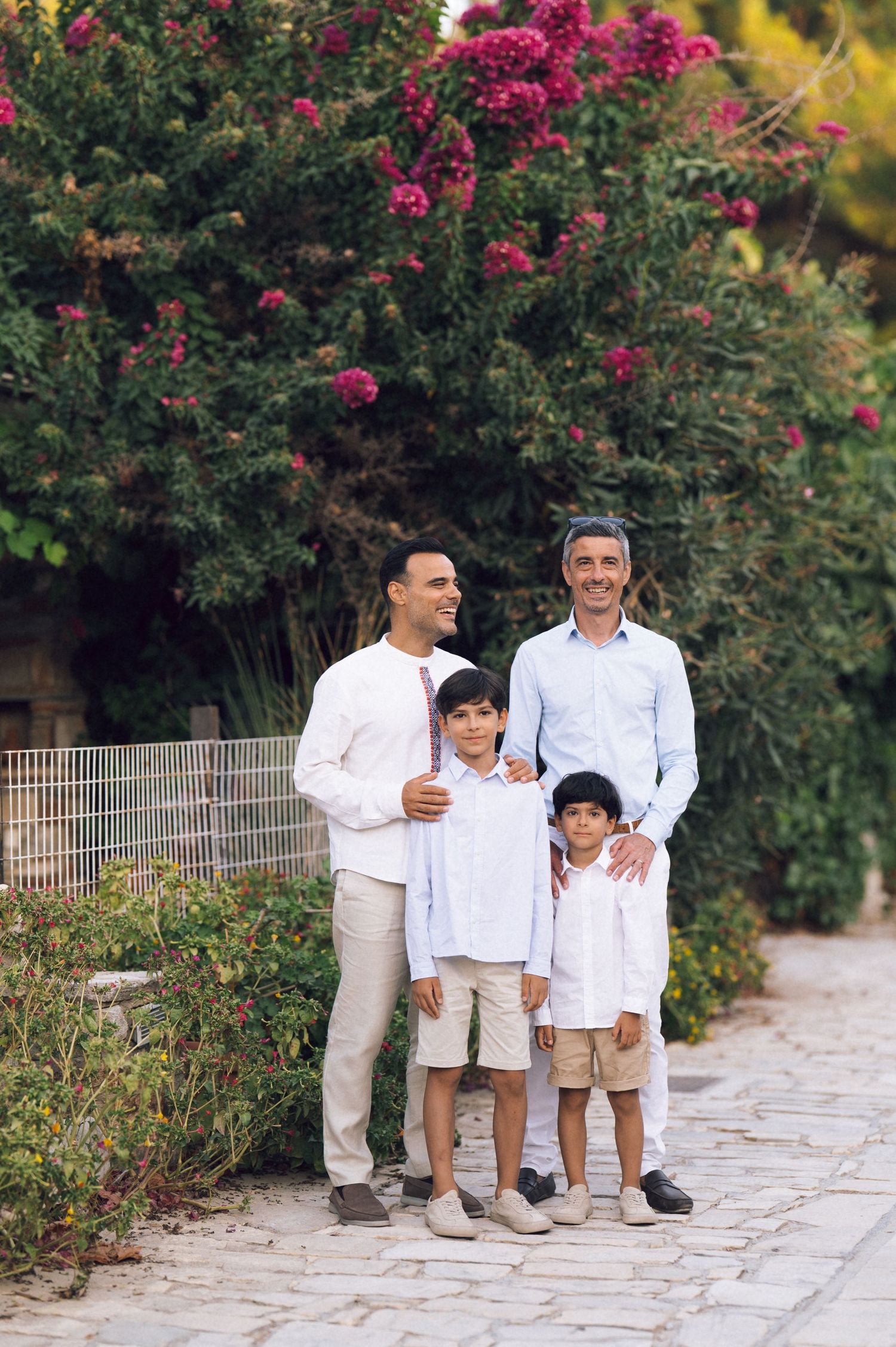Two men and two boys pose together on a stone path surrounded by lush green shrubs and pink flowering trees.