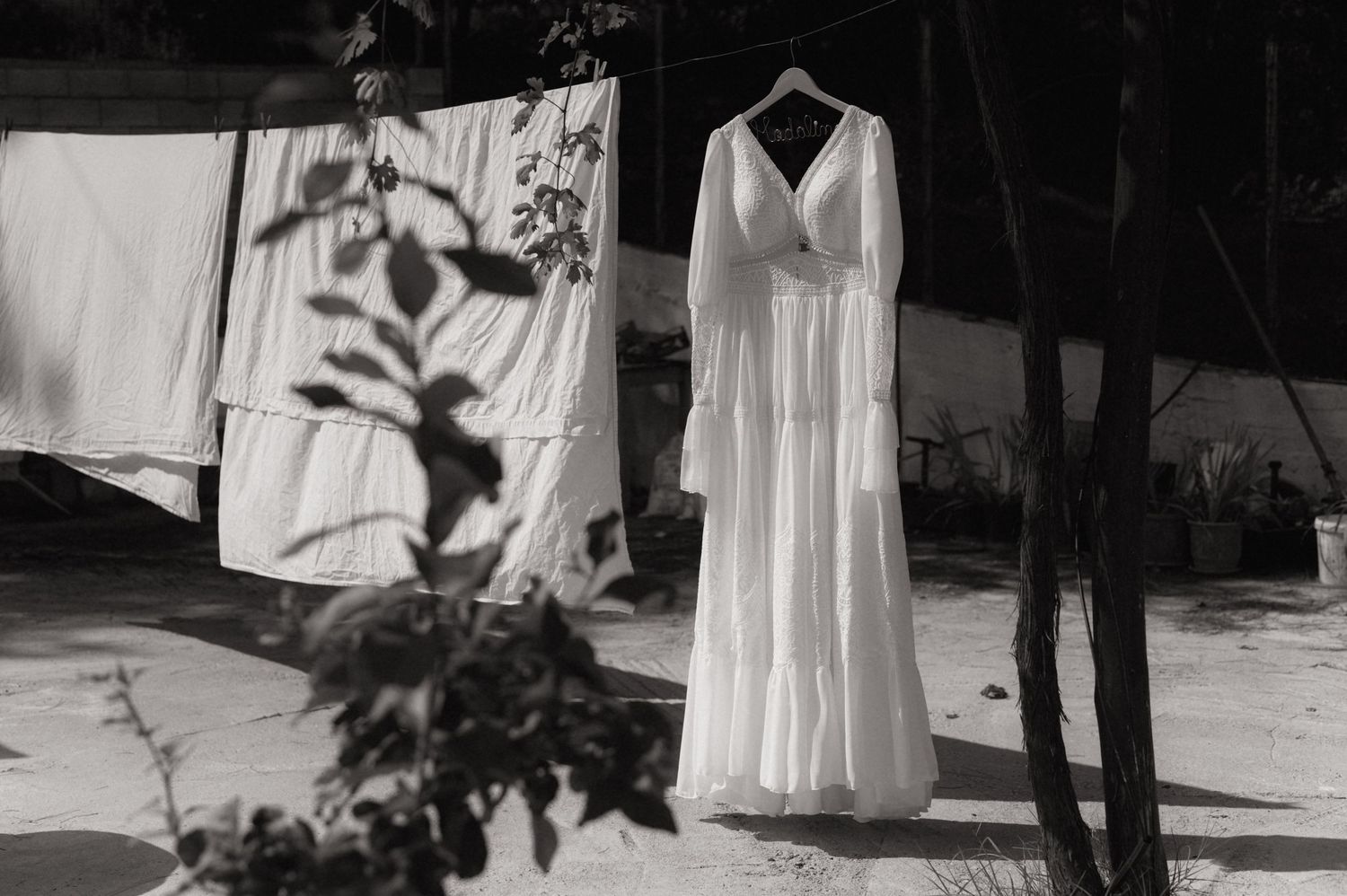 Black and white photo of an elegant white wedding dress hanging on a clothesline outdoors among trees.