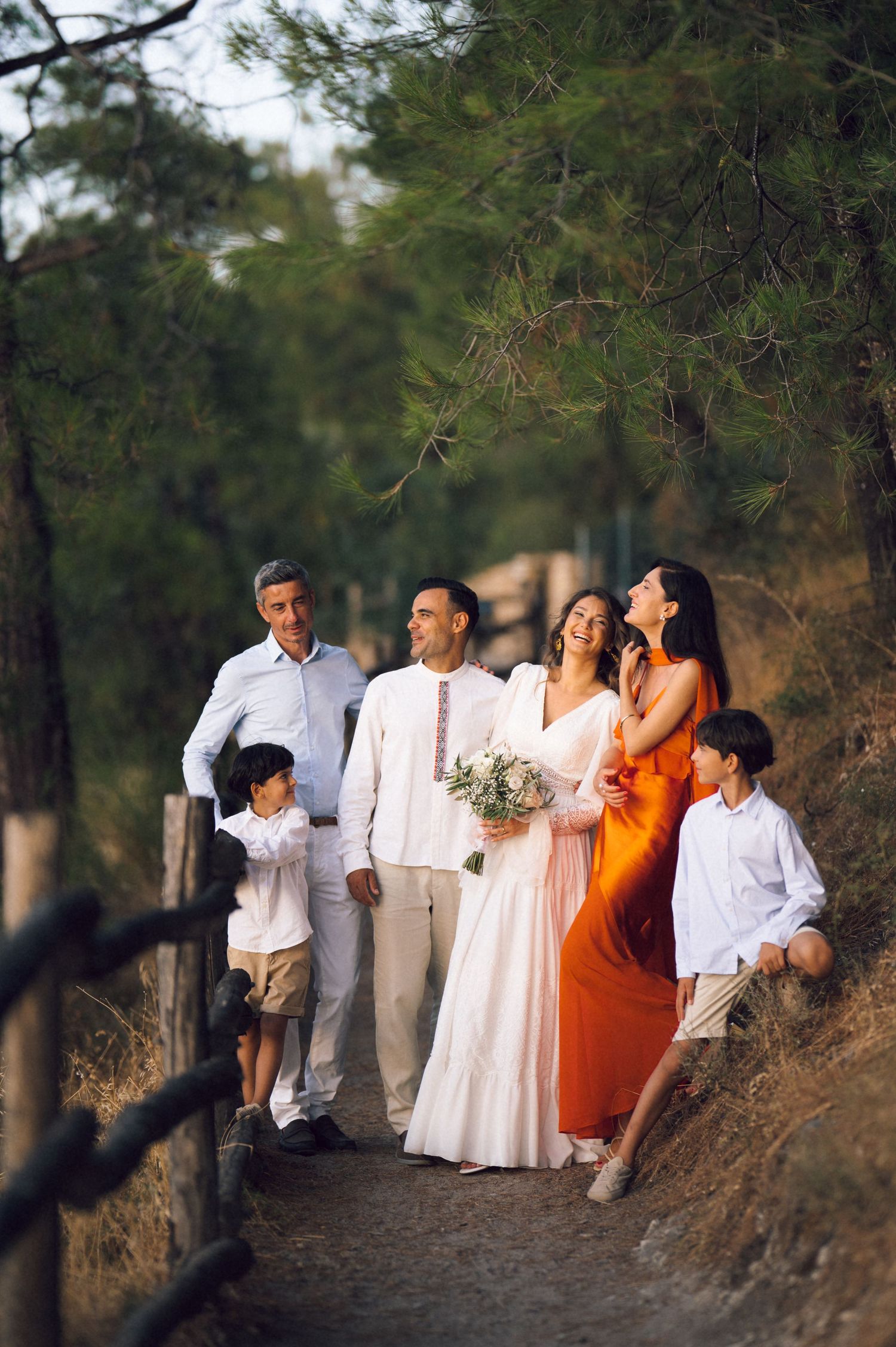 Wedding couple walks forest path with family, bride in white gown, bridesmaid in orange dress, two children accompany them.