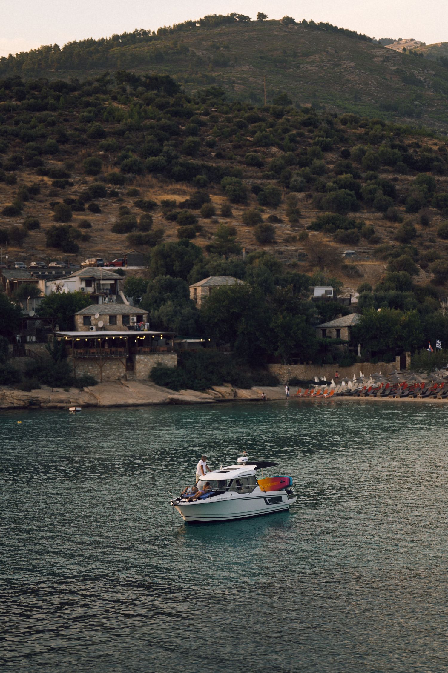A small white motorboat cruises on turquoise waters near a hillside village with lush green trees at dusk.