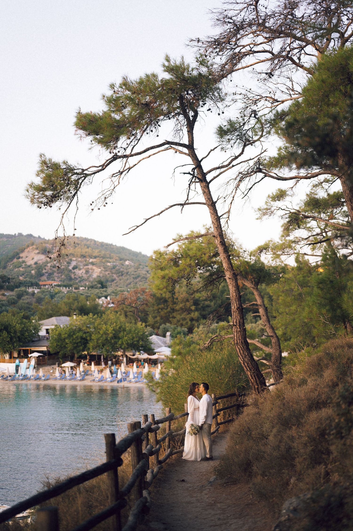 Couple embracing on a scenic waterfront path lined with pine trees and a wooden fence at golden hour.