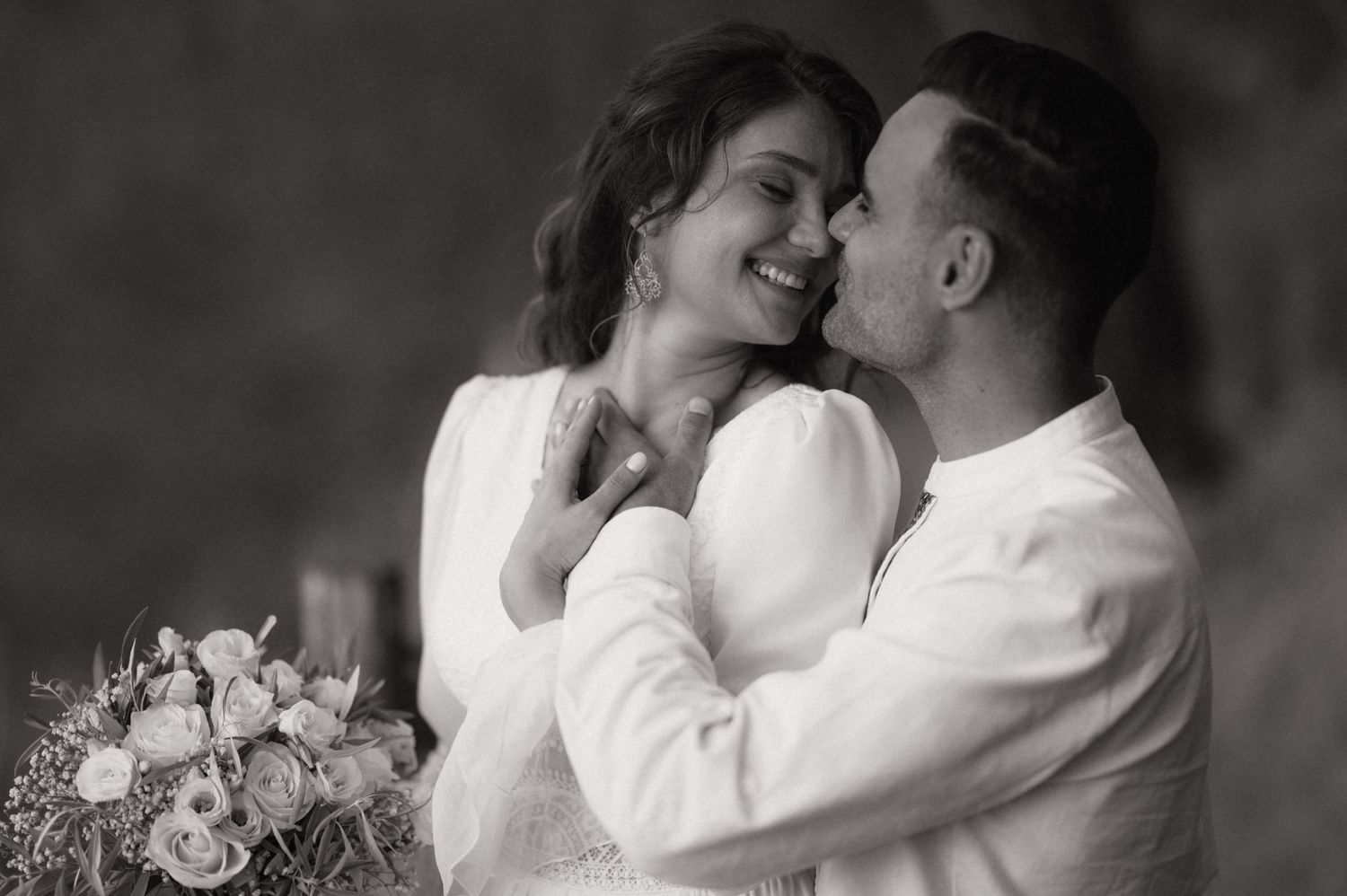 Black and white wedding photo of a smiling bride holding bouquet and groom in white shirt sharing a romantic moment.