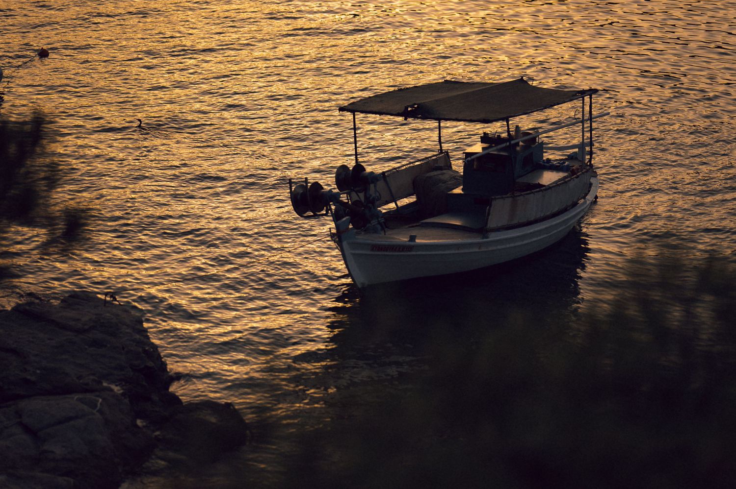 A small motorboat with a canopy anchored near rocky shore at golden sunset, reflecting warm light on calm water.